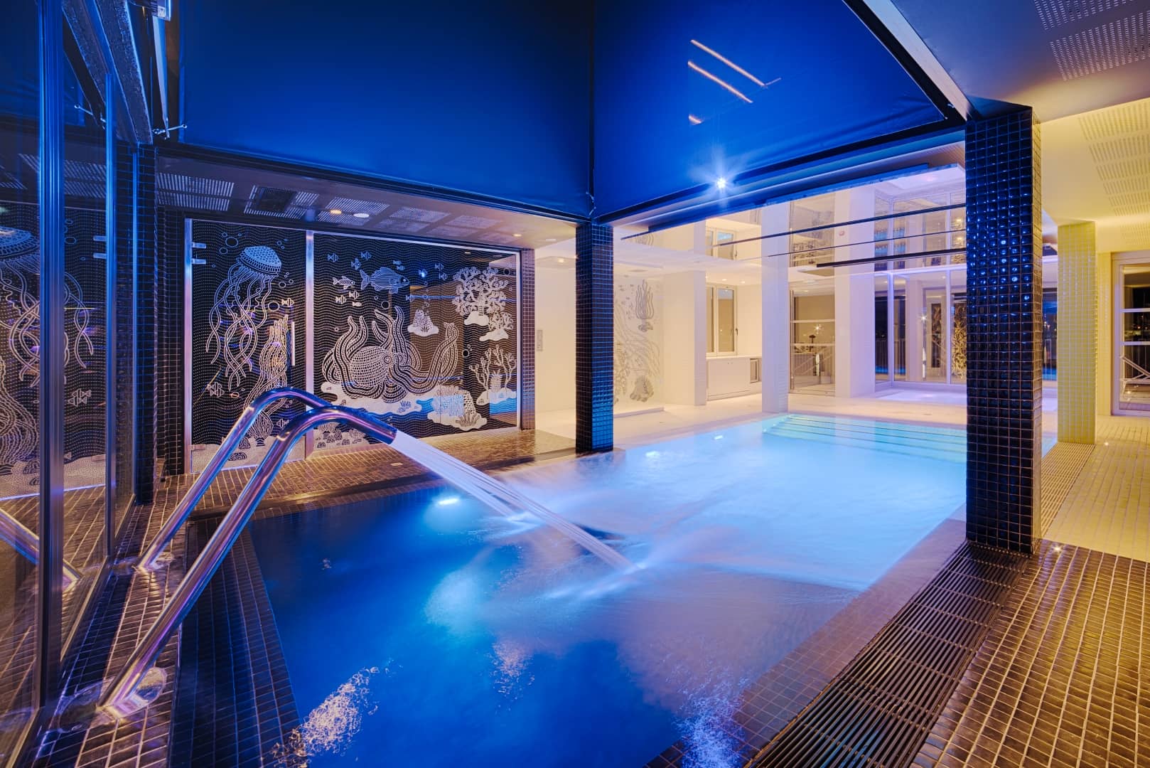 Bassin spa intérieur évoquant une ambiance séminaire bien-être, comprenant une rambarde chromée près d'un jet d'eau éclairé en bleu au premier plan, un mur en mosaïque de créatures marines en noir et blanc au centre, et un espace piscine lumineux avec marches carrelées visible à travers une arche en arrière-plan.