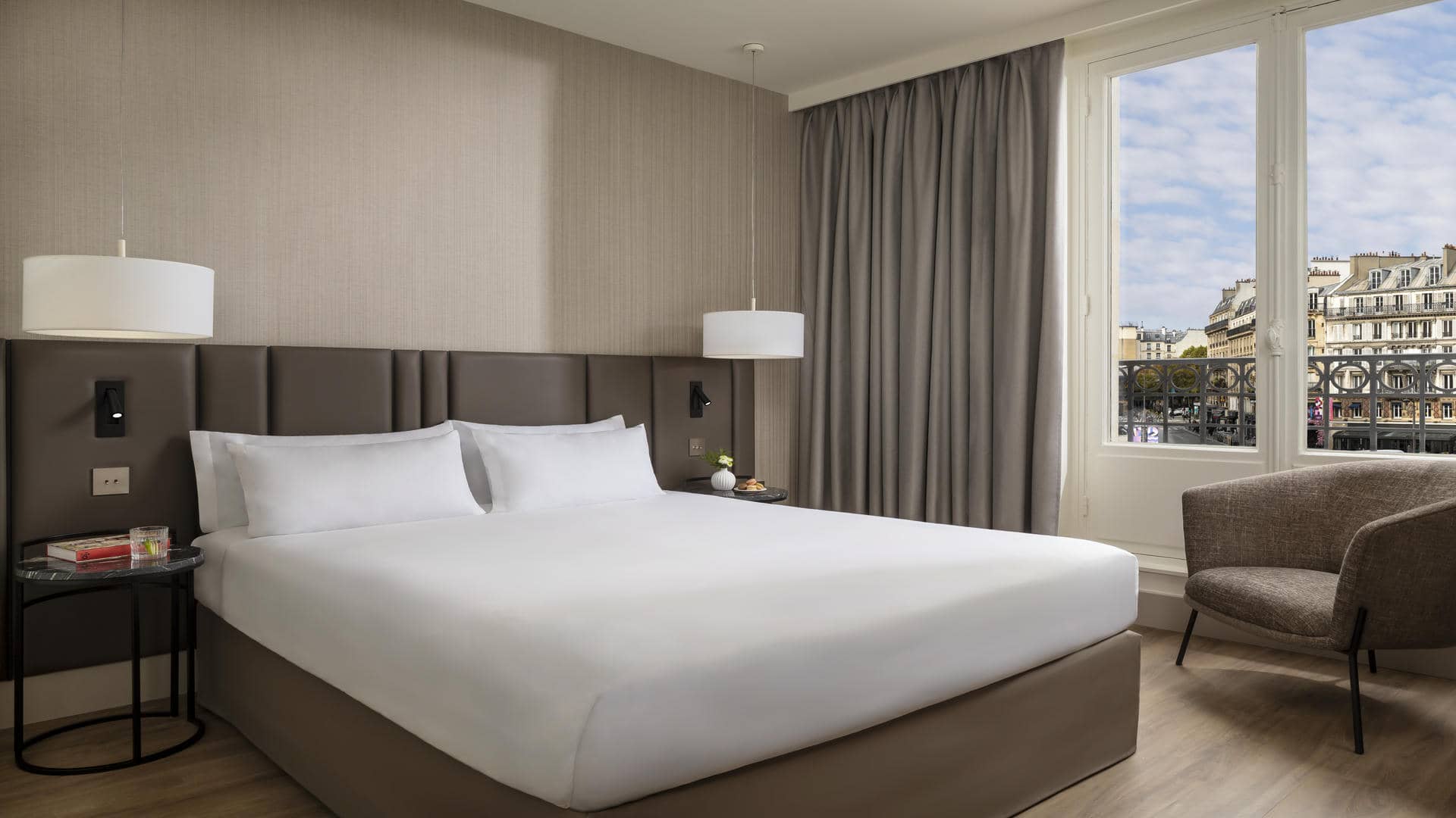 Chambre avec lit double, deux plafonniers blancs descendus au niveau des oreillers, épais rideaux beiges et grande fenêtre haussmannienne avec vue sur le quartier parisien.