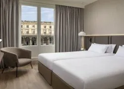 Chambre avec lits twin, tête de lit sur toute la largeur de la pièce, épais rideaux beiges, grandes fenêtres haussmanniennes avec vue sur la gare, et bureau avec une pomme et des livres.