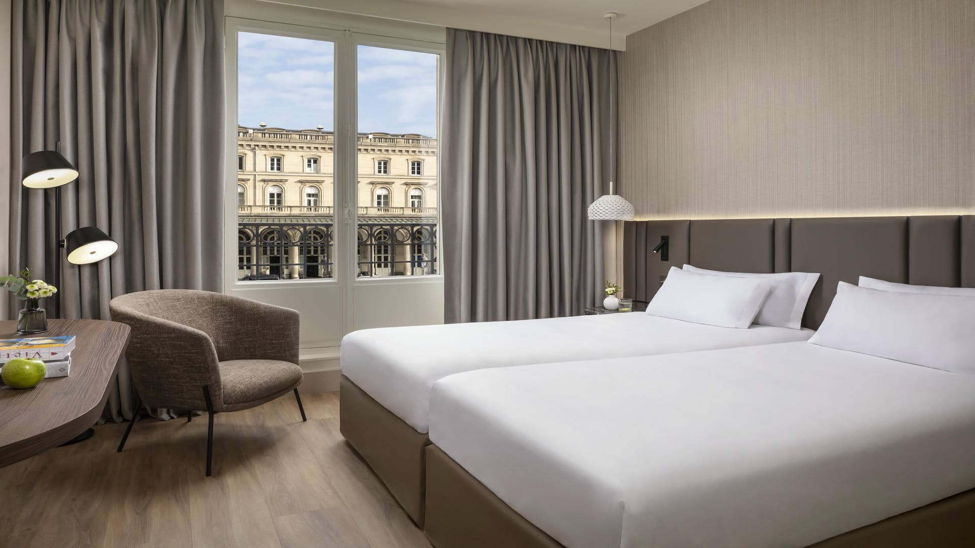 Chambre avec lits twin, tête de lit sur toute la largeur de la pièce, épais rideaux beiges, grandes fenêtres haussmanniennes avec vue sur la gare, et bureau avec une pomme et des livres.