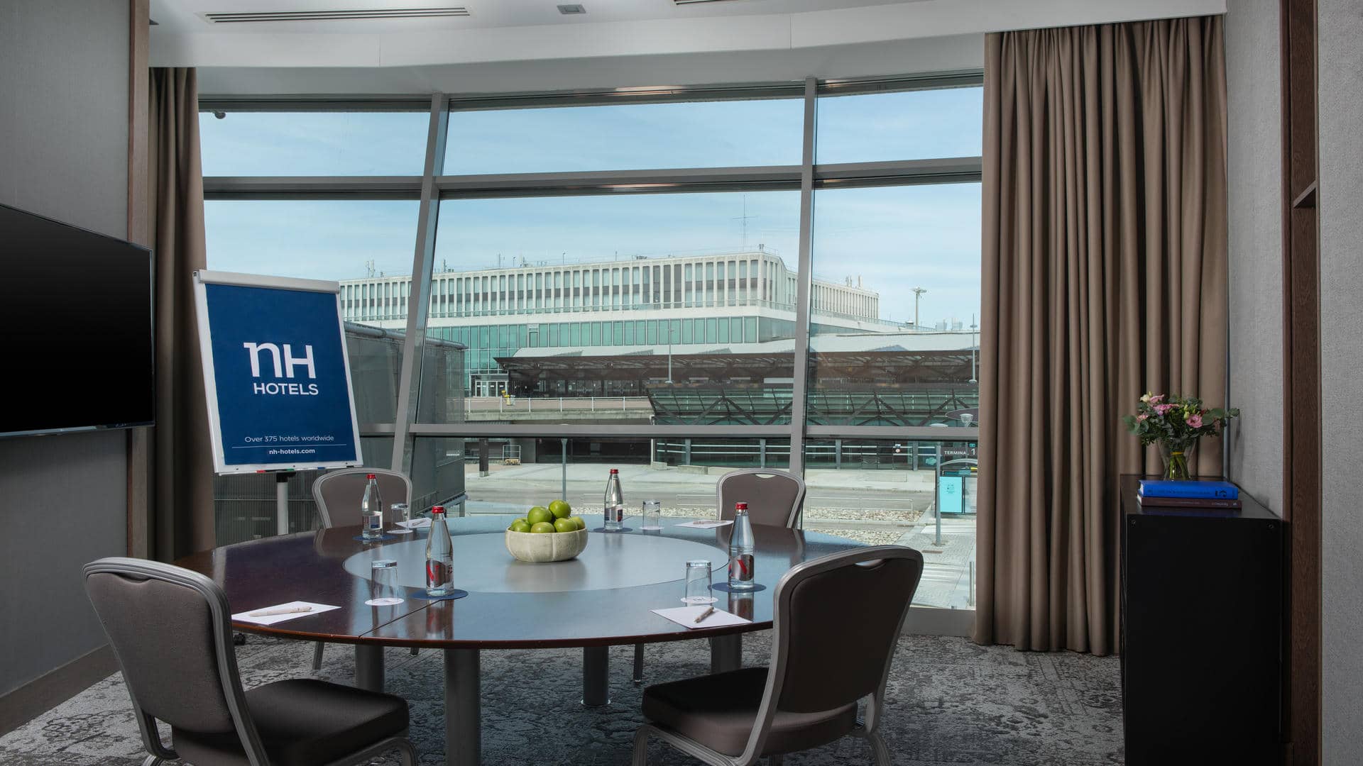 Small training or meeting room with a round wooden table surrounded by four chairs, a wall-mounted TV screen, floor-to-ceiling windows overlooking the airport, and a paper board displaying the NH Hotels logo.