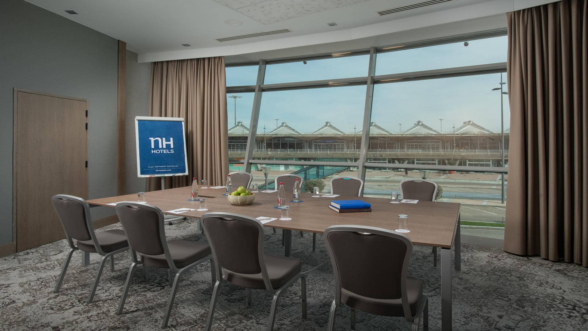 Seminar room with grey carpet and beige curtains partially open onto Lyon Airport. A paper board displaying the NH Hotels logo stands beside a wooden boardroom table arranged to accommodate eight people, seated on either side.