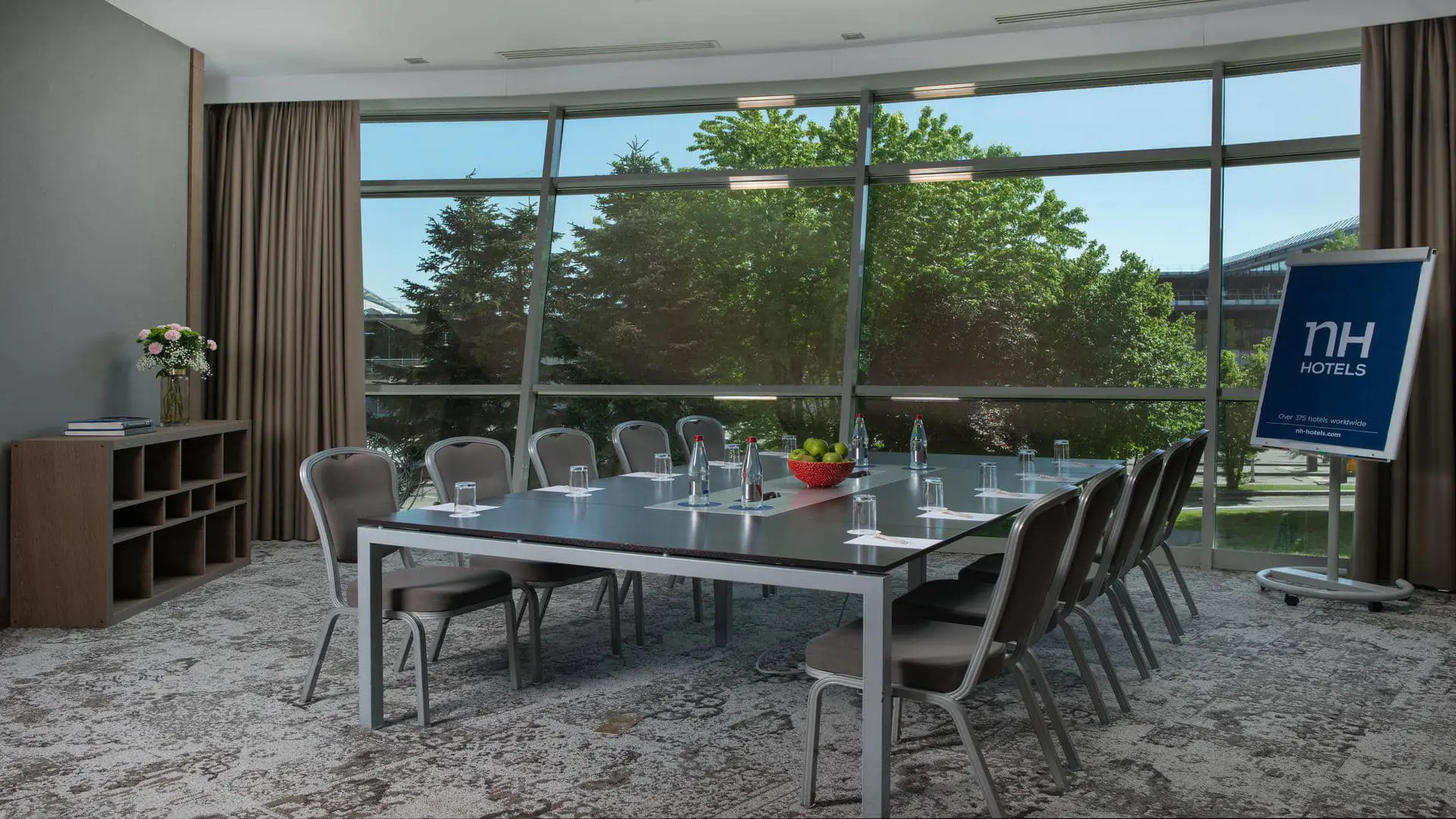 Seminar room with grey carpet and beige curtains partially open onto the surrounding greenery of the property. A paper board displaying the NH Hotels logo stands beside a metal boardroom table arranged to accommodate ten people, seated on either side. A wooden console nearby holds a bouquet of flowers and several books.