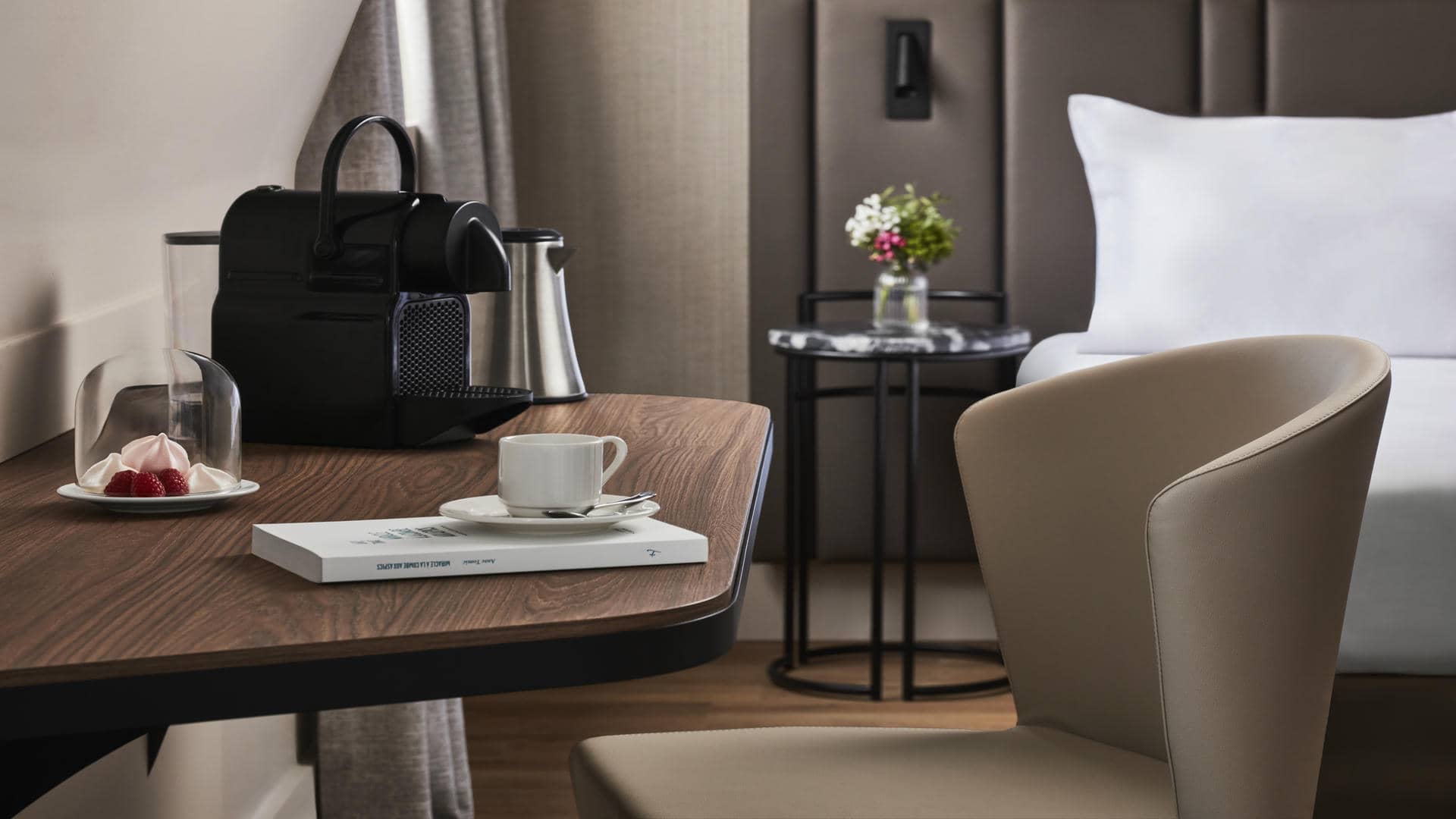 Table en bois foncé fixée au mur beige, avec machine Nespresso, meringues et fraises sous cloche, livre et tasse à café, chaise en tissu devant, et en arrière-plan vue sur le lit double et son chevet fleuri.