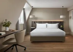 Chambre avec lit double, tête de lit couvrant presque toute la largeur de la pièce, épais rideaux beiges, bureau avec douceurs sucrées, bouquet et livres, chaise de bureau à roulettes, et photo en noir et blanc d’un chef de gare.
