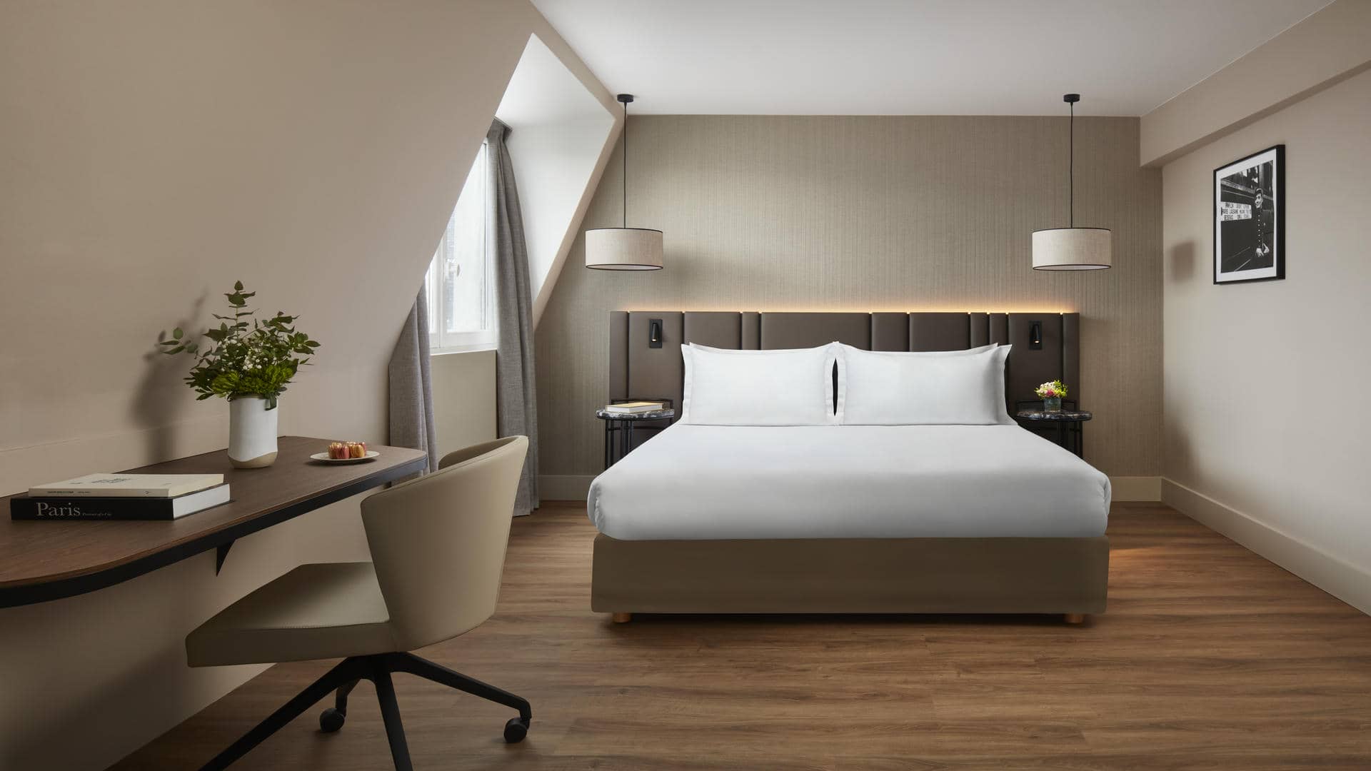 Chambre avec lit double, tête de lit couvrant presque toute la largeur de la pièce, épais rideaux beiges, bureau avec douceurs sucrées, bouquet et livres, chaise de bureau à roulettes, et photo en noir et blanc d’un chef de gare.