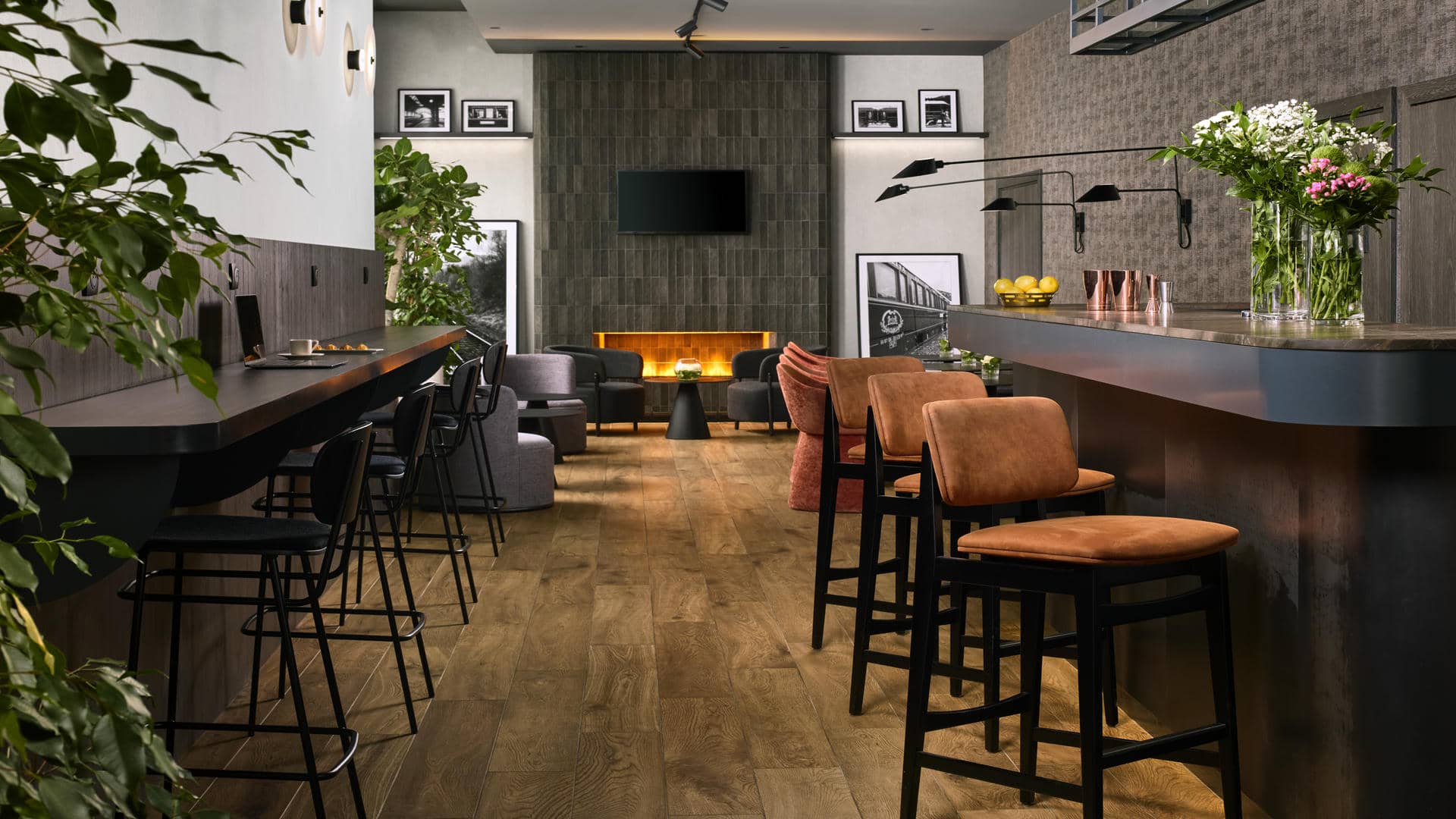 Comptoir de bar avec chaises hautes en tissu terracotta à droite de la salle, tablettes en bois contre le mur en face avec chaises hautes noires pour coworker, cheminée, photographies en noir et blanc et espaces salons.