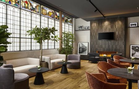 Le bar de l’hôtel séminaire se compose, à droite, de vitraux, de canapés séparés par des plantes, avec tables basses noires devant chaque assise, d’une cheminée et, en face, de fauteuils noirs ou terracotta disposés autour de petites tables basses noires.