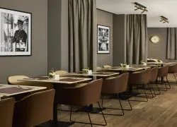 Tables beiges pour deux personnes avec chaises marron, dressées le long du mur gris de la salle petit-déjeuner, décoré de photographies en noir et blanc illustrant la vie en gare.