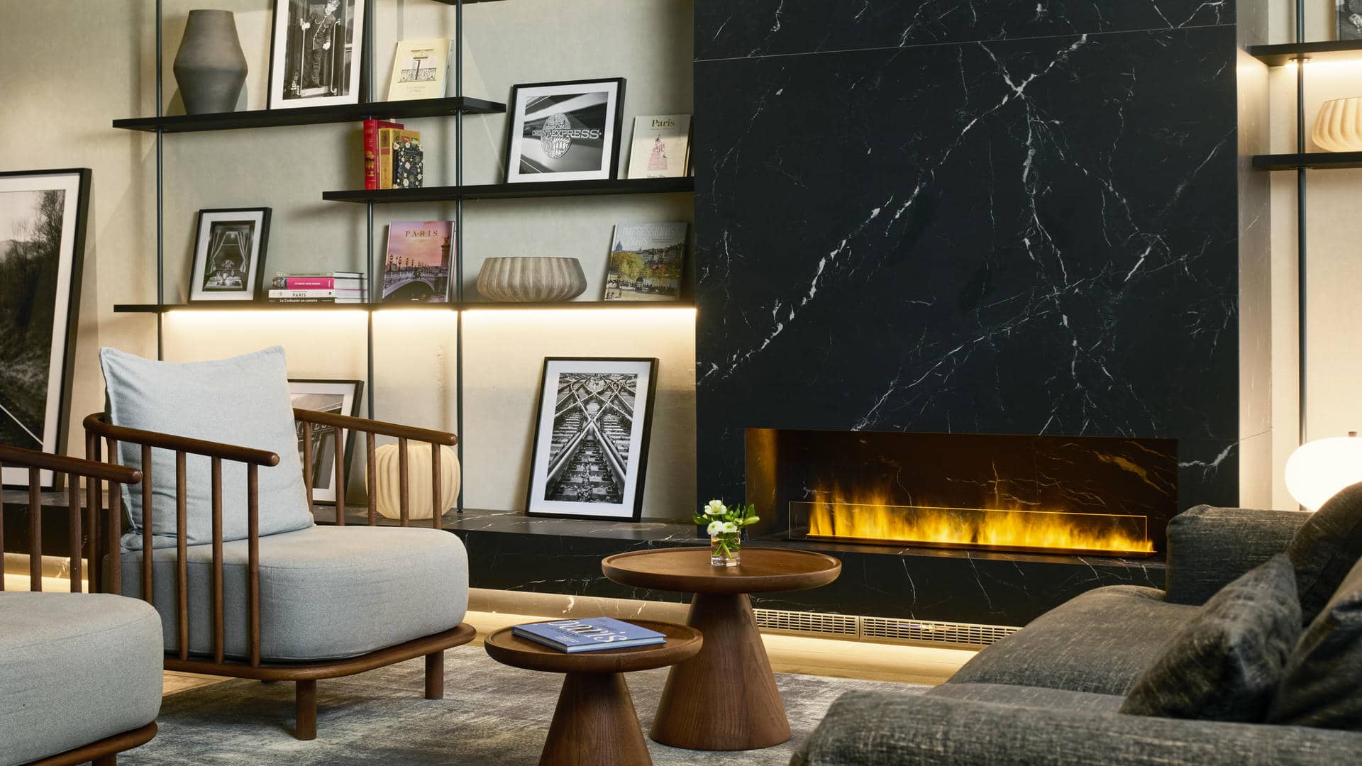 Le bar de l’hôtel séminaire se compose d’une cheminée entourée d’étagères décorées, de fauteuils gris devant de petites tables en bois et d’un grand canapé gris.