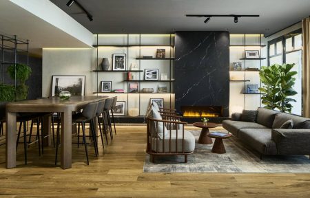 Salon avec parquet, étagère murale noire décorée de photographies en noir et blanc de la Gare de l’Est, cheminée centrale, canapé gris et fauteuils autour de petites tables basses, grande table d’hôtes avec chaises de bar face à une autre étagère murale noire ornée de plantes.