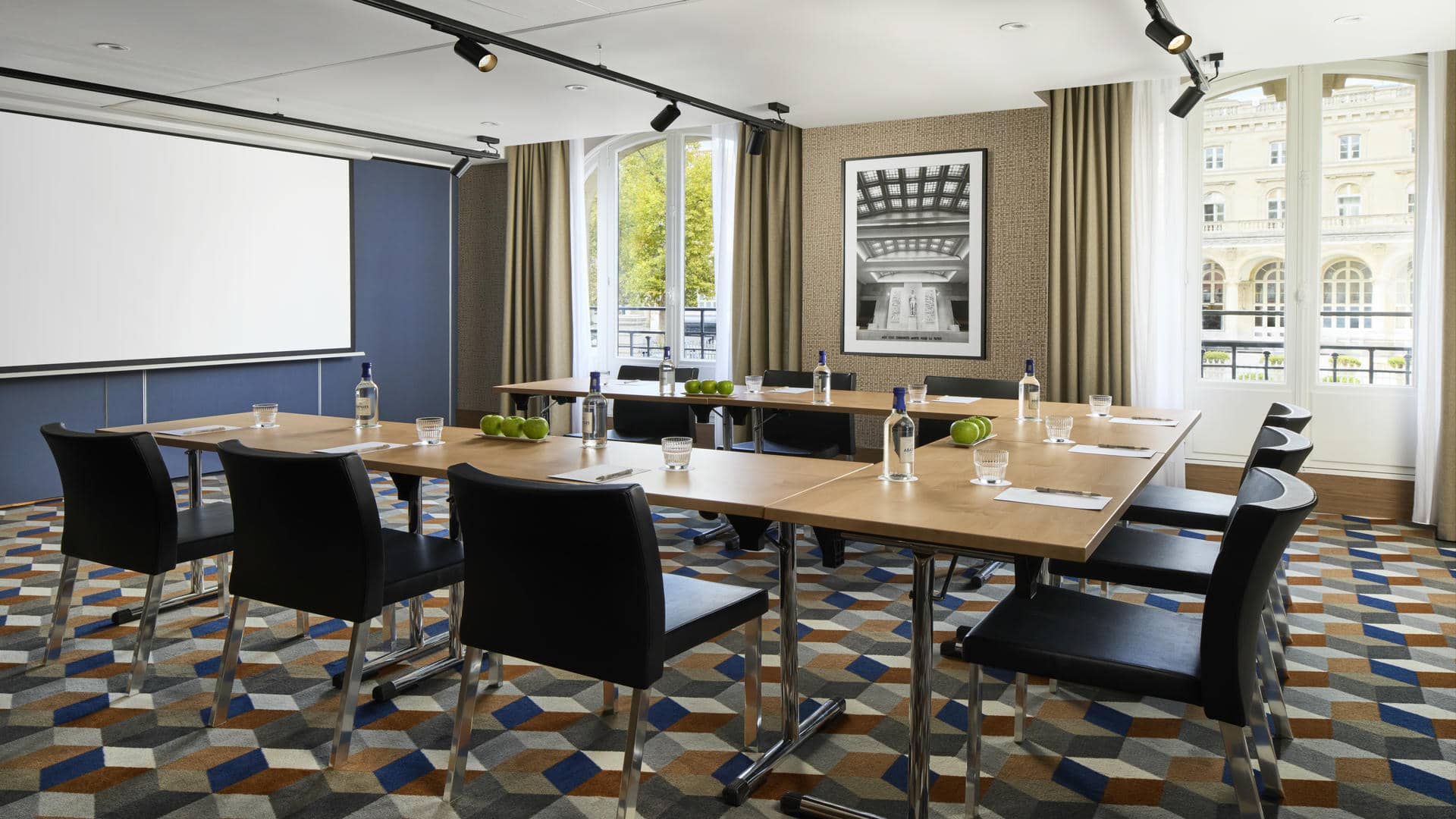 Salle de réunion avec six tables en bois, chaises noires, moquette à motifs cylindriques, mur tendu beige, photo en noir et blanc de la Gare de l’Est, grandes fenêtres donnant sur la gare, écran mural.
