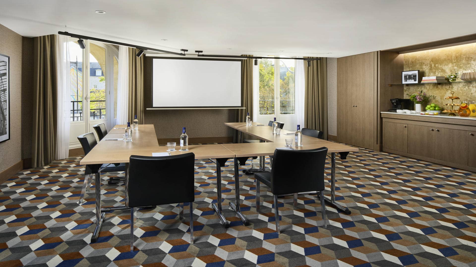 Salle de réunion avec six tables en bois, chaises noires, moquette à motifs cylindriques, mur tendu beige, photo en noir et blanc de la Gare de l’Est, grandes fenêtres donnant sur la gare, écran mural et meuble aménagé pour une pause séminaire.