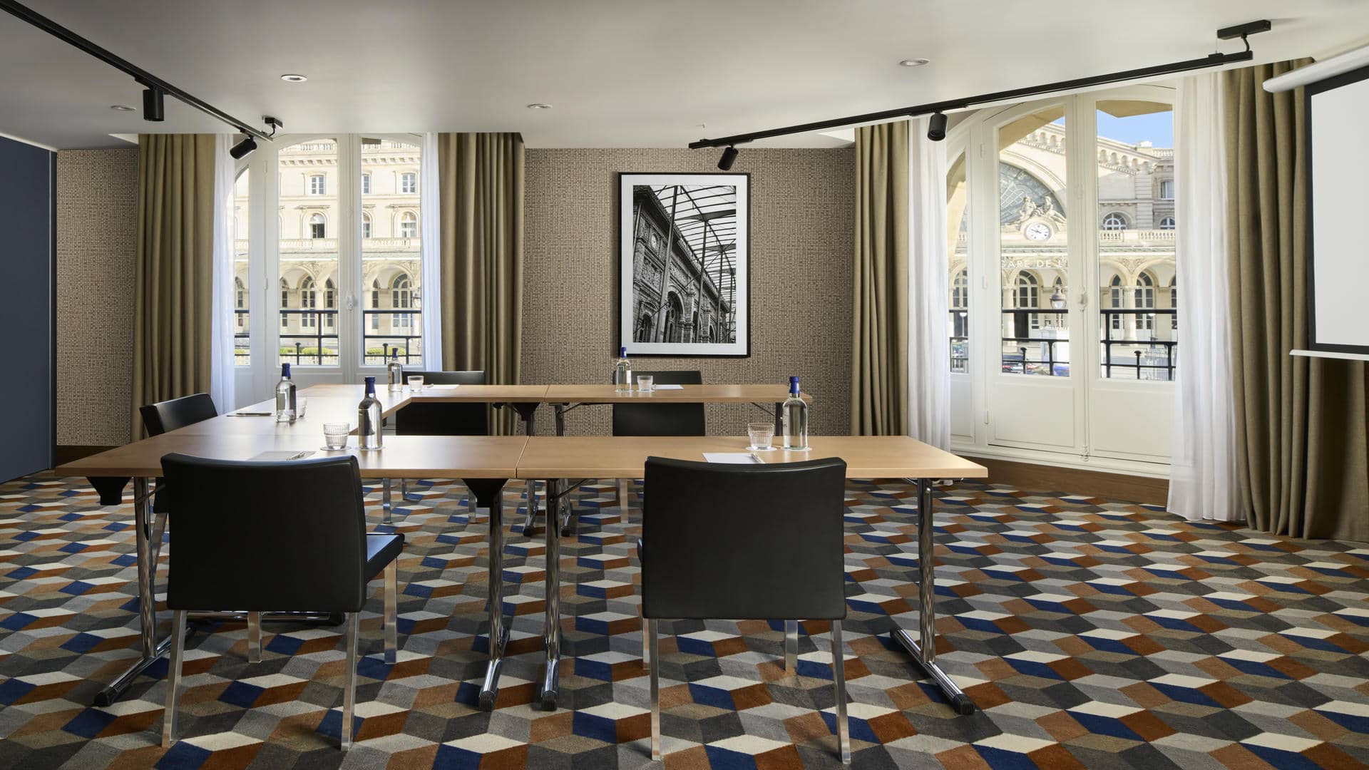 Salle de réunion avec quatre tables en bois, chaises noires, moquette à motifs cylindriques, mur tendu beige, photo en noir et blanc de la Gare de l’Est et grandes fenêtres donnant sur la gare.