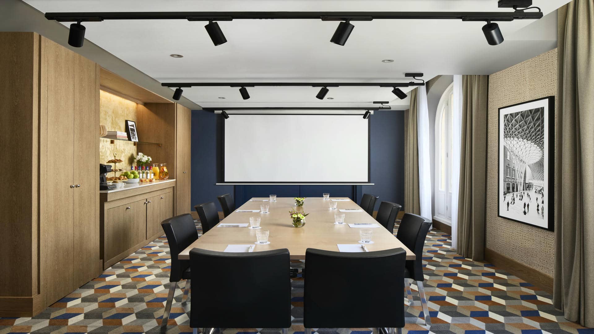 Salle de réunion avec tables en bois réunies en pavé, cloisons bleu roi, revêtement mural tissé beige, écran mural et grande photo murale.