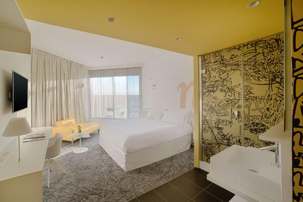 Chambre d'hôtel pour hébergement groupe en vue générale avec un lit blanc au centre, un bureau blanc et une chaise avec une télévision murale à gauche, un lavabo blanc au premier plan à droite, un canapé jaune et une table basse blanche près de la fenêtre à l'arrière-plan, et une cloison à motifs graphiques noirs.