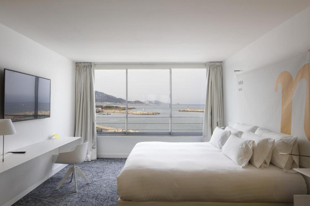 Chambre d'hôtel pour séminaire en bord de mer, avec un grand lit blanc à droite au premier plan, un bureau blanc et une télévision murale à gauche, et une grande baie vitrée donnant sur la mer, un port de plaisance et des collines en arrière-plan, sur une moquette grise à motifs.