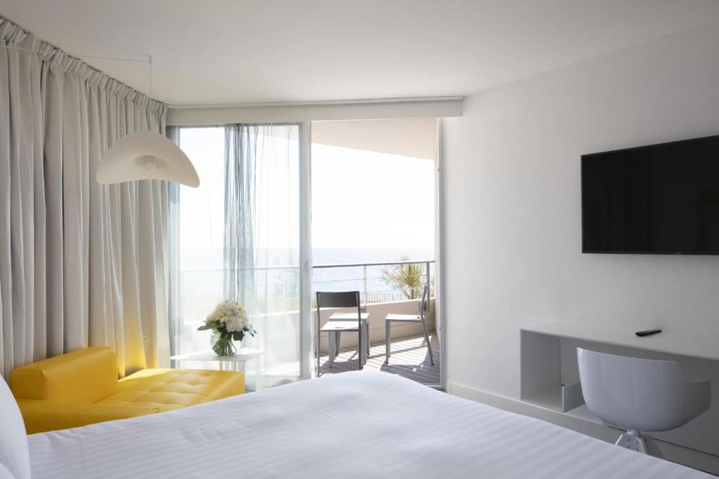 Chambre d'hôtel pour séminaire en bord de mer, avec un grand lit blanc au premier plan, une méridienne jaune et un lampadaire blanc à gauche, une télévision murale avec un bureau blanc à droite, et un balcon avec deux chaises donnant sur la mer en arrière-plan.