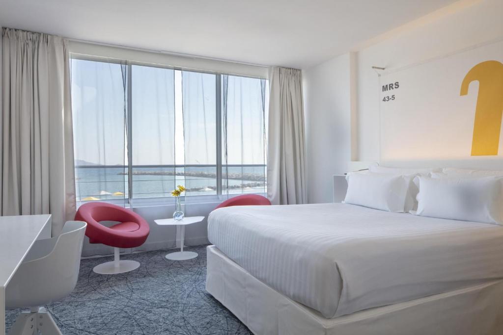 Chambre d'hôtel pour séminaire en bord de mer, avec un grand lit blanc à droite au premier plan, un bureau blanc à gauche, deux fauteuils rouges et une table d'appoint ronde blanche devant une grande baie vitrée au centre, et une vue sur la mer en arrière-plan sur une moquette grise à motifs.