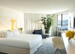 Chambre d'hôtel propice à un séjour en séminaire en bord de mer, comprenant un lit blanc avec logo « n » jaune au-dessus de la tête de lit au premier plan, une chaise longue bleu marine et une grande plante verte au centre, et un balcon avec baies vitrées coulissantes donnant sur le port de plaisance et les montagnes en arrière-plan.