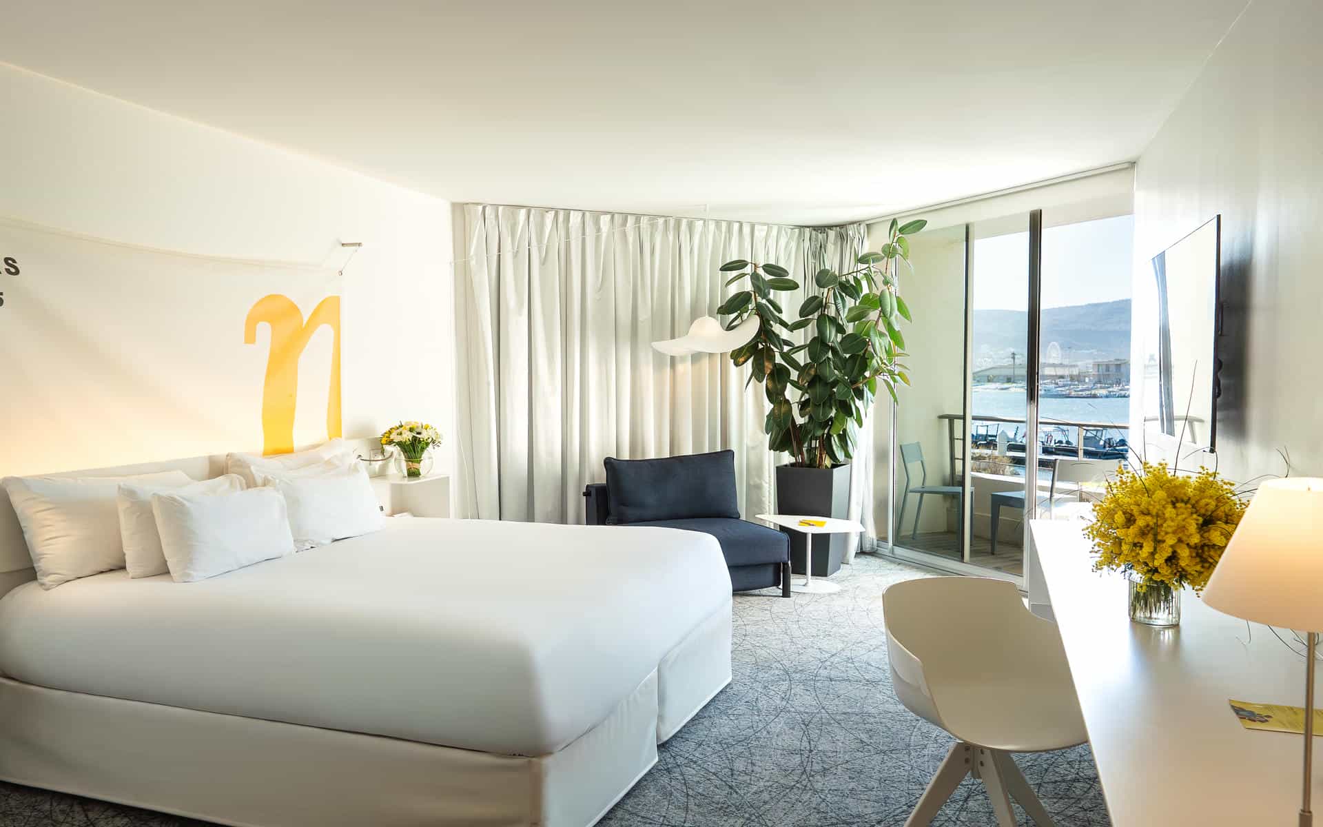 Chambre d'hôtel propice à un séjour en séminaire en bord de mer, comprenant un lit blanc avec logo « n » jaune au-dessus de la tête de lit au premier plan, une chaise longue bleu marine et une grande plante verte au centre, et un balcon avec baies vitrées coulissantes donnant sur le port de plaisance et les montagnes en arrière-plan.
