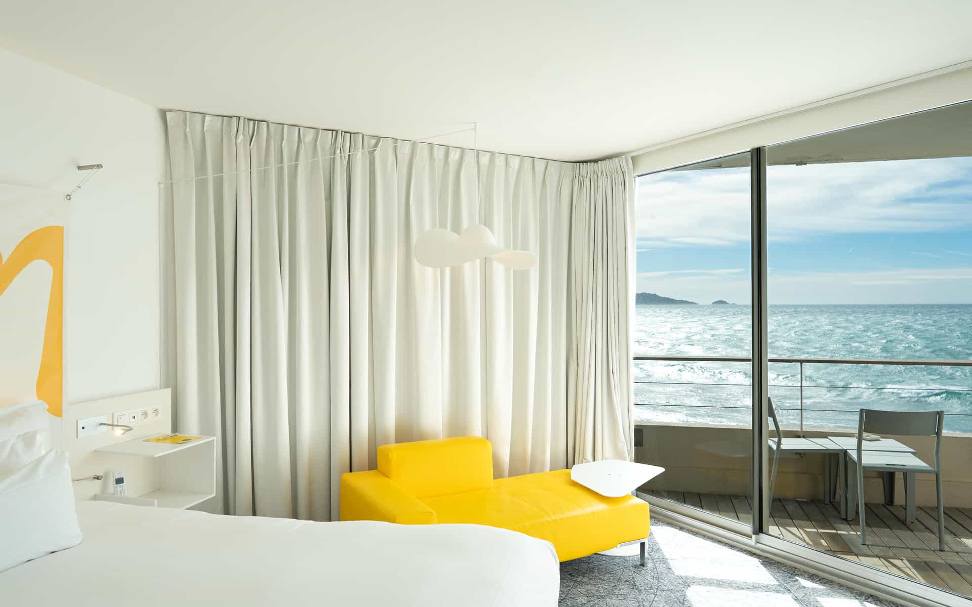 Chambre d'hôtel pour séminaire en bord de mer avec un lit blanc au premier plan à gauche, une chauffeuse jaune accompagnée d'une table d'appoint blanche au centre, des rideaux blancs toute hauteur, une grande baie vitrée coulissante ouvrant sur un balcon avec une table et des chaises à droite, et la mer à l'arrière-plan.