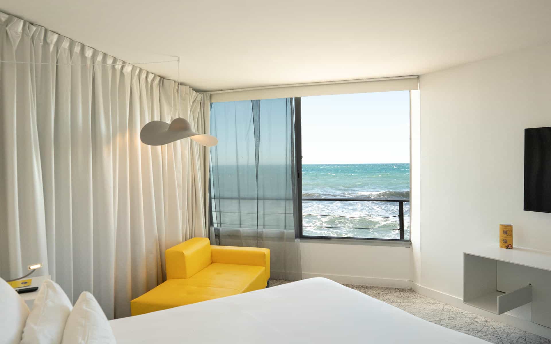 Chambre d'hôtel pour séminaire au vert avec un lit blanc au premier plan, une chauffeuse jaune à gauche, des rideaux blancs toute hauteur, une grande fenêtre avec vue sur la mer au centre, et une télévision murale au-dessus d'un meuble blanc à droite.