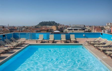 Hotel rooftop swimming pool with beige sun loungers lined up on both sides and along the back edge, glass railings and a blue-tiled pool wall surrounding the water, a metal pool ladder on the right, and a hillside city with a wooded hilltop and rooftops visible under a clear sky in the background, a panoramic rooftop pool for a wellness seminar.