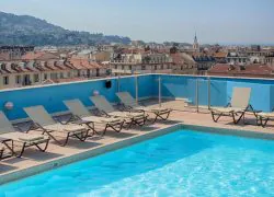 Piscine sur le toit d'un hôtel avec chaises longues beiges alignées le long du bord au premier plan, garde-corps vitrés et paroi de piscine carrelée bleue bordant l'eau, marquage de profondeur « 1,0 m » sur le carrelage de la piscine, et toits de Nice avec colline et clocher visibles sous un ciel voilé en arrière-plan, une piscine panoramique sur le toit pour un séminaire bien-être.