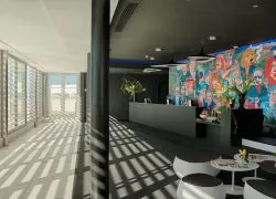 Espace réception d'hôtel propice à une arrivée en séminaire en bord de mer, comprenant des tabourets ronds noirs et blancs avec petites tables et tulipes jaunes au premier plan, un comptoir d'accueil sombre avec suspensions lumineuses au centre, et une grande fresque colorée de personnages illustrés sur le mur en arrière-plan, avec un couloir baigné de lumière aux fenêtres à claire-voie à gauche.