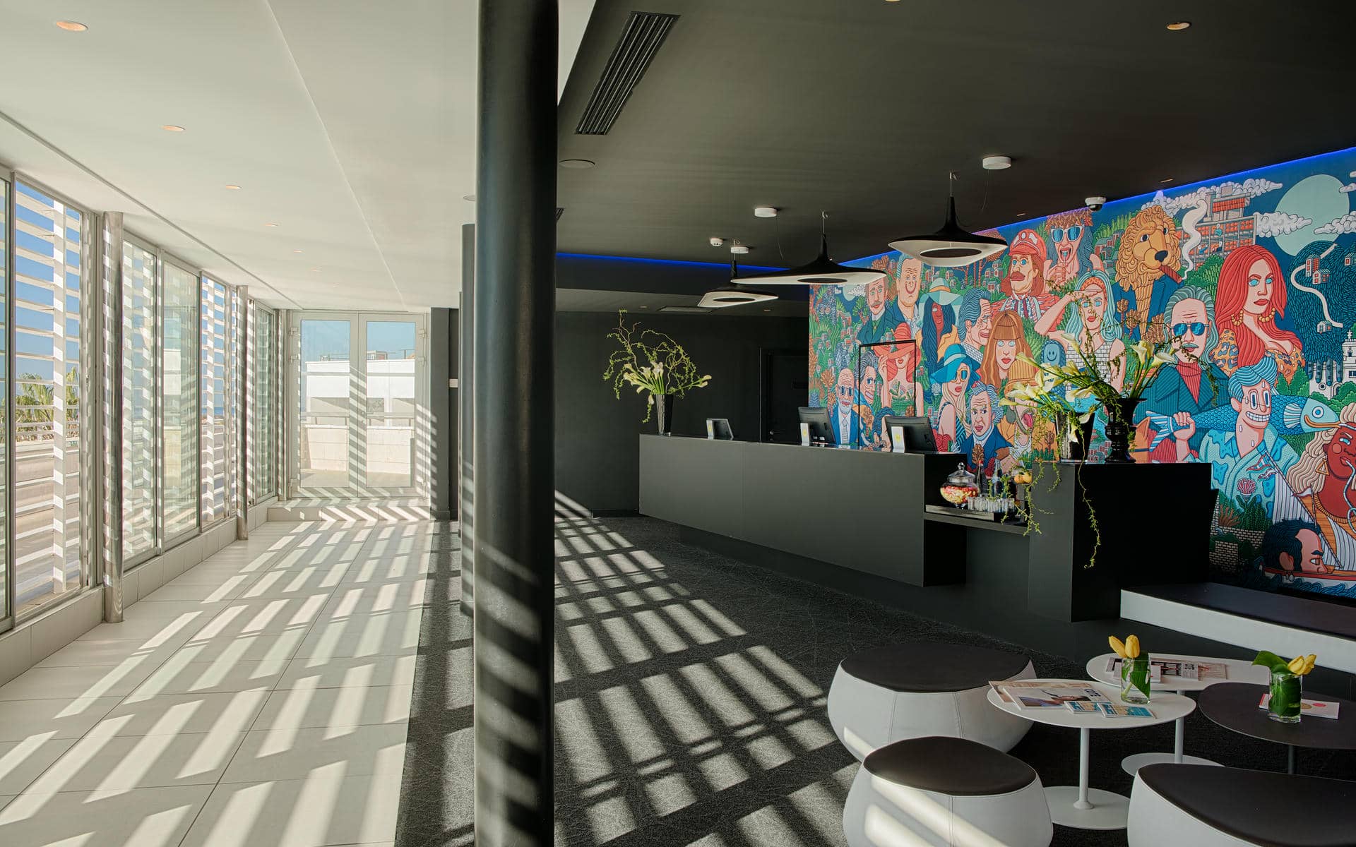 Espace réception d'hôtel propice à une arrivée en séminaire en bord de mer, comprenant des tabourets ronds noirs et blancs avec petites tables et tulipes jaunes au premier plan, un comptoir d'accueil sombre avec suspensions lumineuses au centre, et une grande fresque colorée de personnages illustrés sur le mur en arrière-plan, avec un couloir baigné de lumière aux fenêtres à claire-voie à gauche.