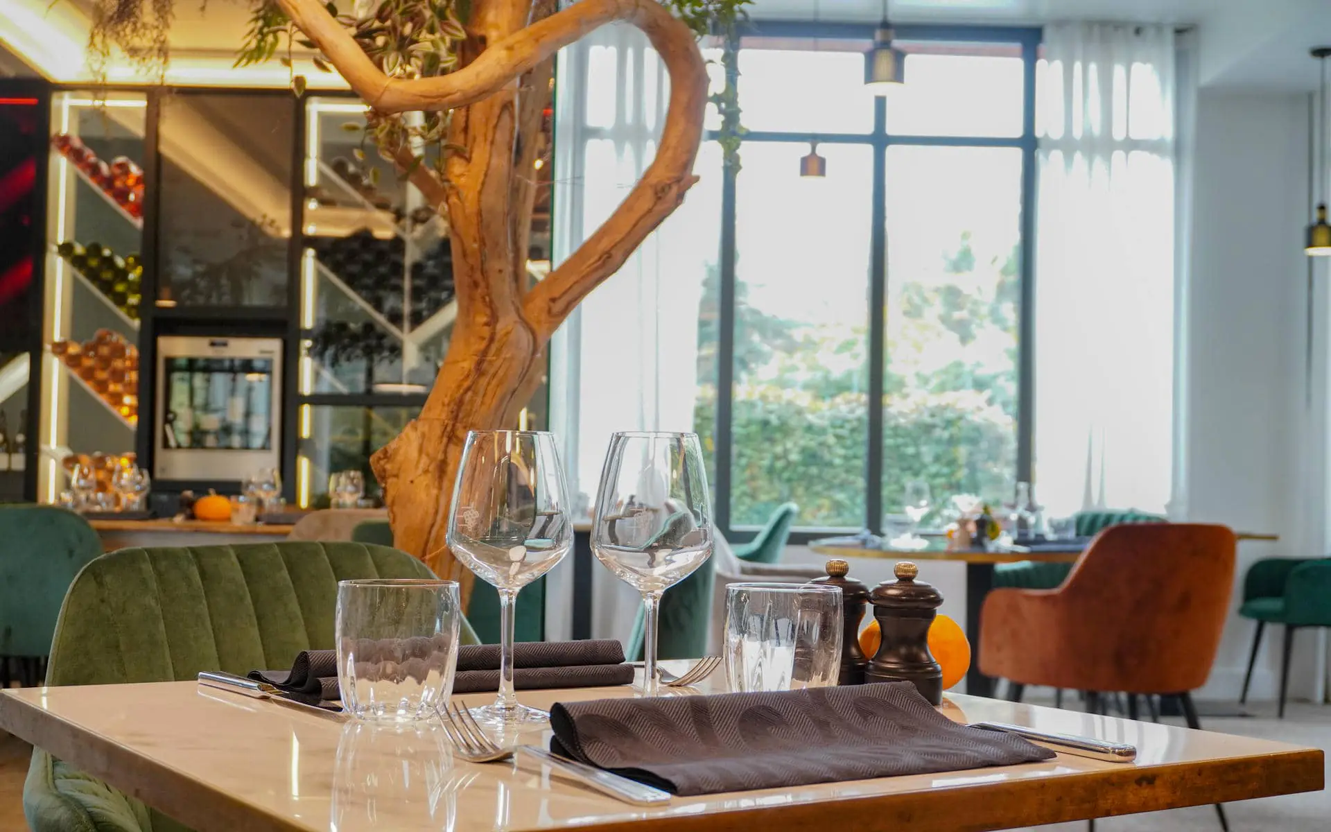 Restaurant table set for on-site catering with wine glasses, water tumblers, dark folded napkins and pepper mills in the foreground, a green velvet banquette on the left, a tree trunk sculpture rising centrally in front of tall windows overlooking greenery, and an illuminated wine rack with bottles visible in the background.