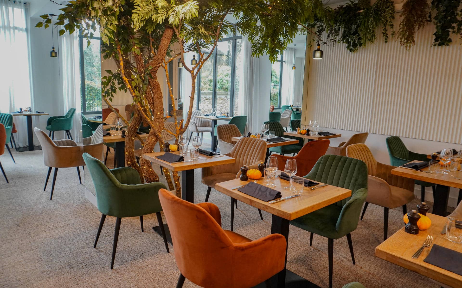 Restaurant dining room for on-site catering with wooden square tables set with wine glasses, dark napkins and small orange pumpkins in the foreground, green, orange and beige velvet chairs surrounding them, an illuminated artificial tree with fairy lights rising through the centre, and tall windows revealing greenery outside in the background.