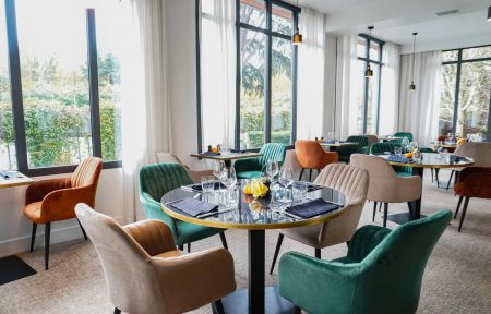 Hotel restaurant for on-site catering, with a round table set with glasses, plates and black napkins in the foreground, velvet armchairs in green, beige and terracotta around the dining area, and large black-framed windows with white curtains overlooking trees in the background.