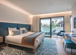 Chambre d'hôtel pour hébergement groupe, avec un lit double doté d'une tête de lit bleue et de coussins sur la gauche, un tapis bleu à motifs et un bureau avec tabouret sur la droite, et une baie vitrée coulissante ouvrant sur un balcon avec des chaises en arrière-plan.