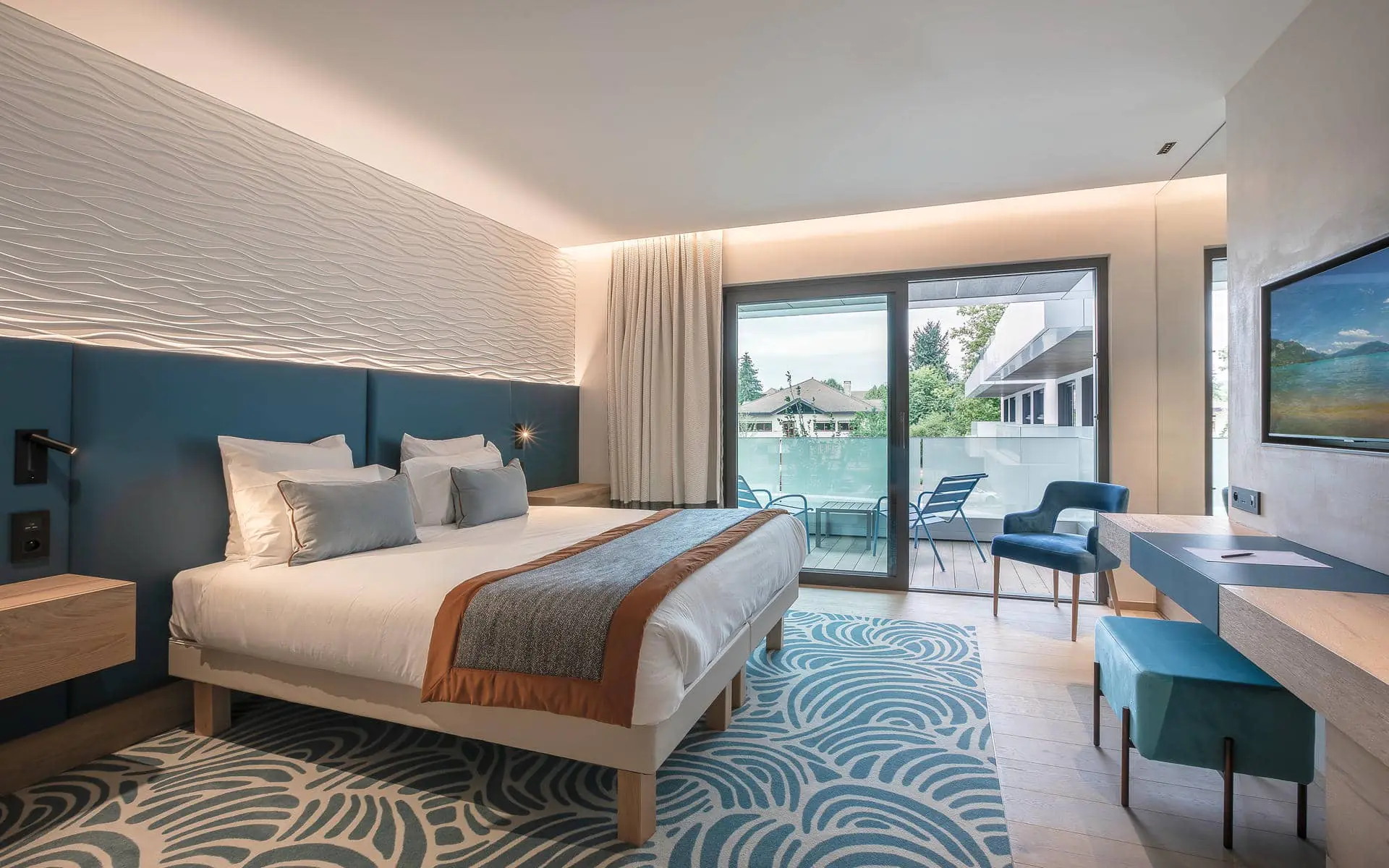 Hotel guest room for group accommodation, with a double bed featuring a blue upholstered headboard and cushions on the left, a blue patterned rug and desk with stool on the right, and a sliding glass door opening onto a balcony with chairs in the background.