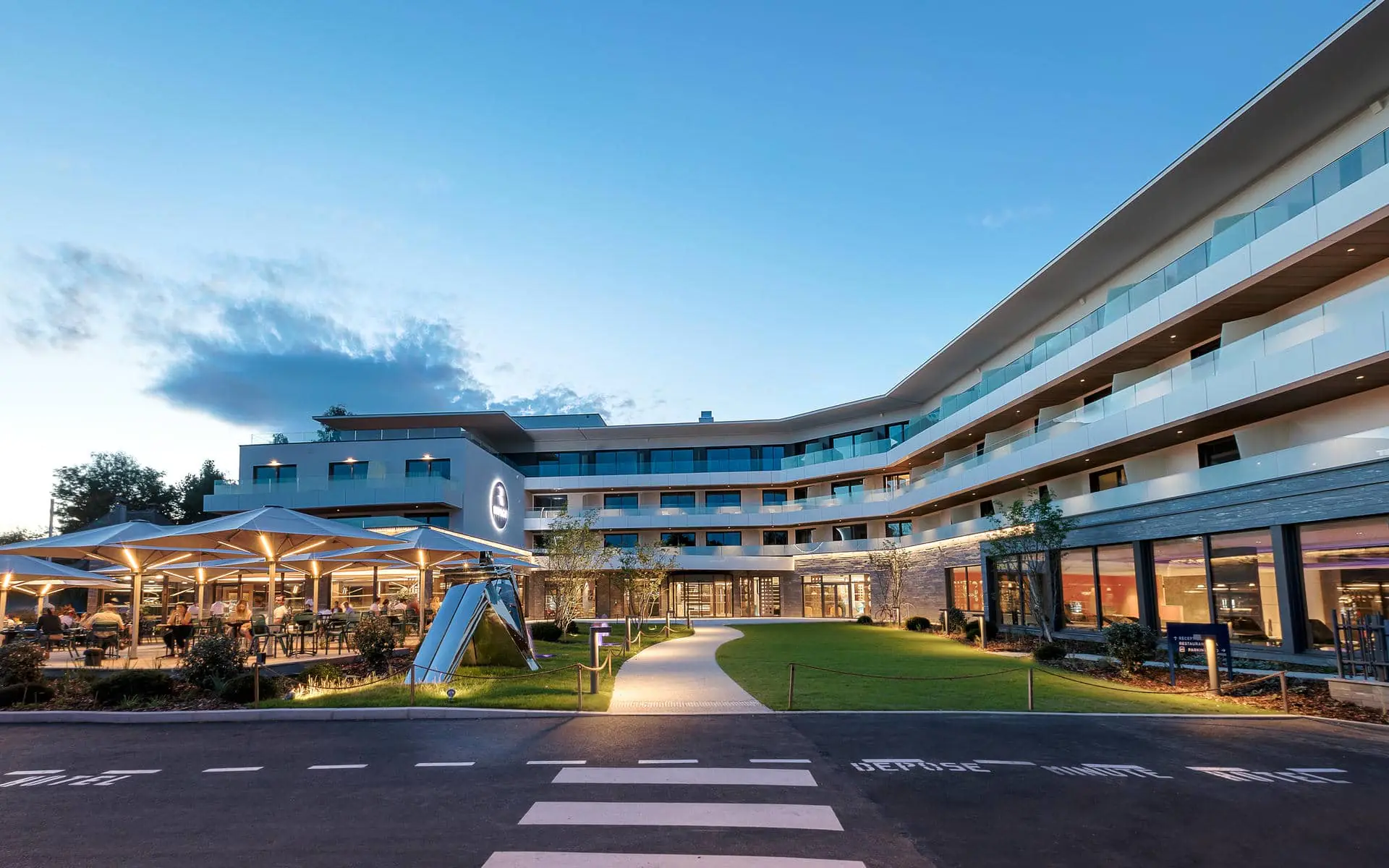 General view of a modern hotel for meeting room hire, featuring a pedestrian crossing and roadway in the foreground, a curved pathway crossing a landscaped lawn in the centre, a terrace with parasols and outdoor tables on the left, and a multi-storey building with glass balconies in the background under a blue evening sky.