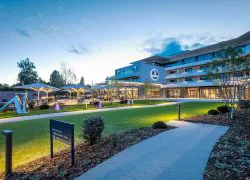 Vue générale d'un hôtel avec jardin paysager pour un séminaire au vert, comprenant une allée revêtue et des massifs plantés au premier plan, des pelouses éclairées avec de petites fontaines au centre, une terrasse avec parasols et tables en extérieur à gauche, et un bâtiment moderne à plusieurs étages avec balcons en arrière-plan.
