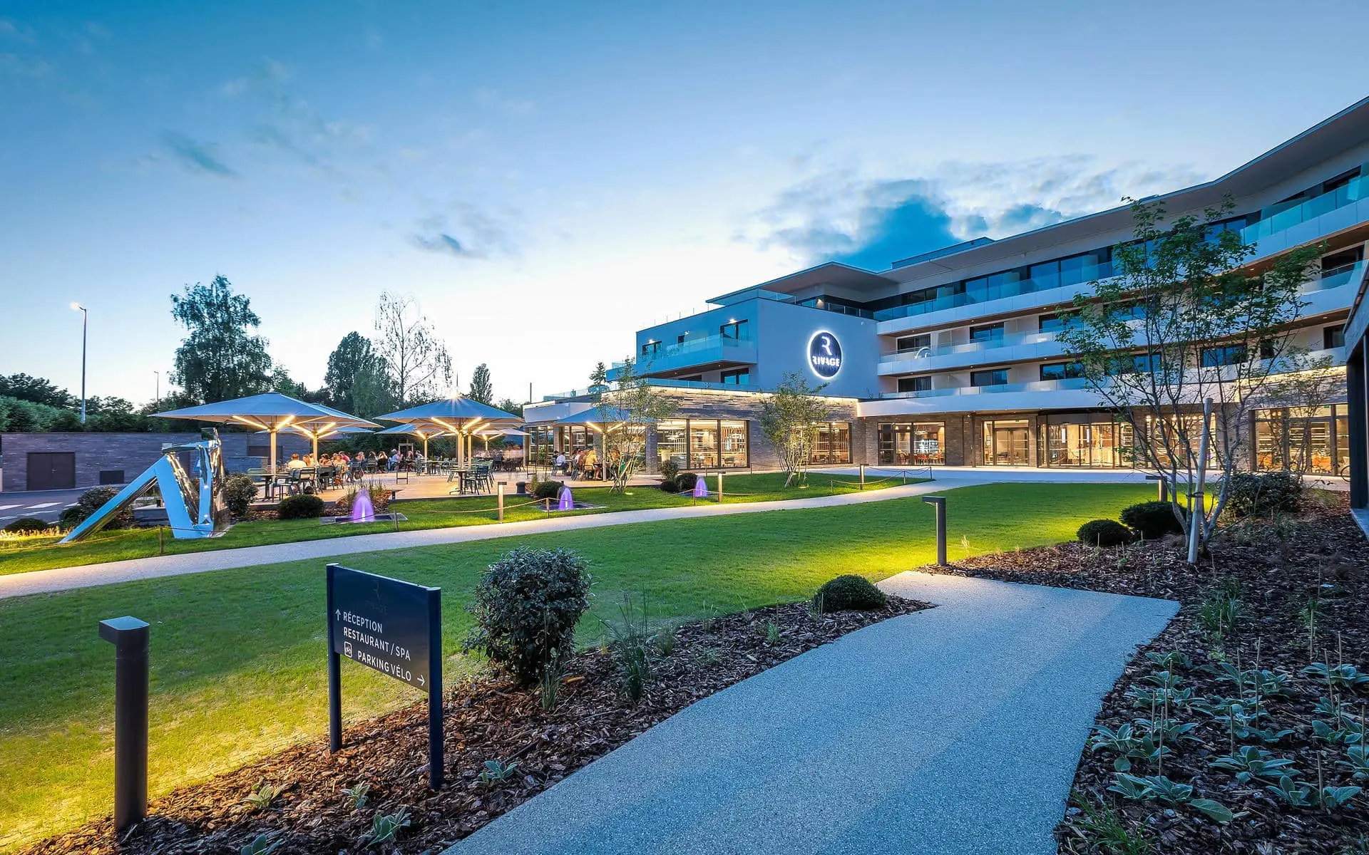 General view of a hotel with a landscaped garden, featuring a paved pathway and planted flower beds in the foreground, illuminated lawns with small fountains in the centre, a terrace with parasols and outdoor tables on the left, and a modern multi-storey building with balconies in the background for a green seminar.