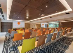 Vue générale d'une salle de formation d'hôtel, comprenant des rangées de chaises multicolores au premier plan, deux écrans de projection et un pupitre à l'avant, une moquette à motifs sous les sièges, et de grandes baies vitrées donnant sur un espace arboré à droite.
