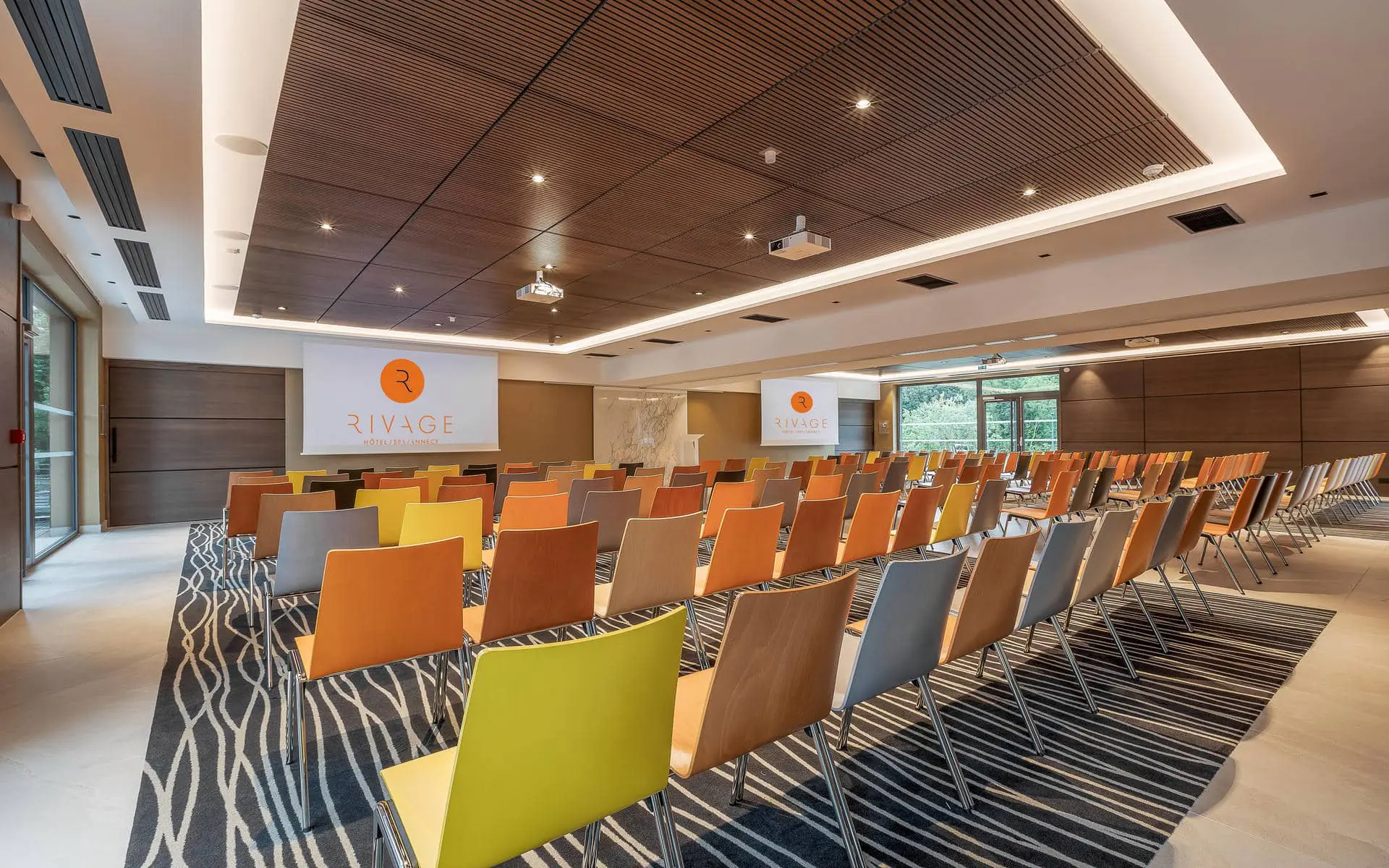 General view of a hotel training room, featuring rows of multicoloured chairs in the foreground, two projection screens and a lectern at the front, patterned carpet beneath the seating, and large windows overlooking greenery on the right.