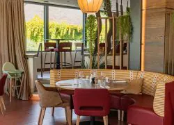 Espace de restauration d'hôtel pour restauration sur place, avec une table ronde dressée de verres et couverts au premier plan, des chaises rouges et une banquette arrondie à rayures au centre, des claustras en bois avec plantes suspendues à droite, et de grandes baies vitrées avec tables hautes et tabourets en arrière-plan.