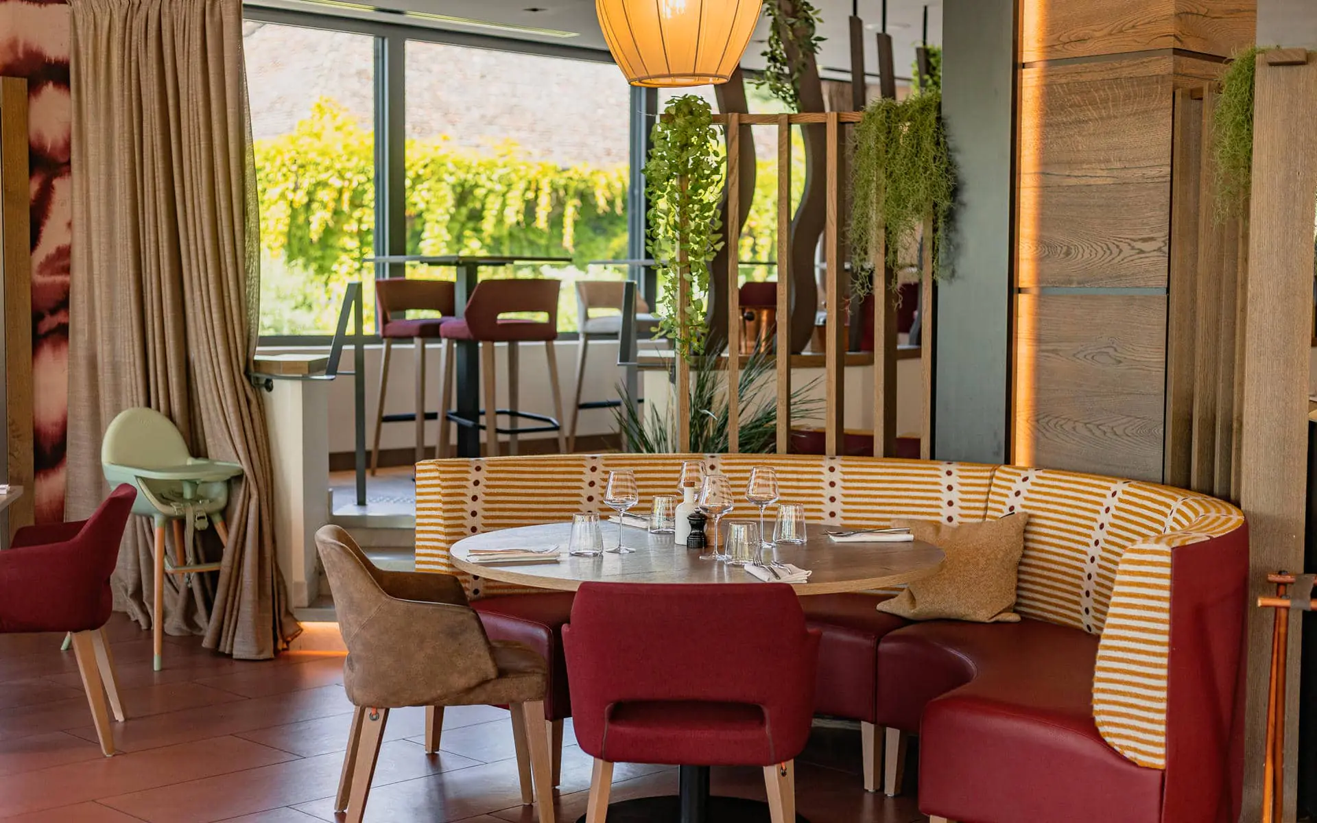 Hotel restaurant dining area for on-site catering, with a round table set with glasses and cutlery in the foreground, red chairs and a curved striped banquette in the centre, wooden partitions with hanging plants on the right, and large windows with high tables and stools in the background.