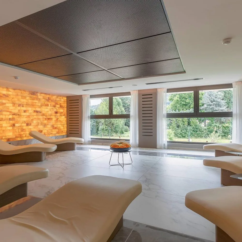 General view of a hotel relaxation room for a wellness seminar, featuring curved cream loungers in the foreground, a small round table in the centre, a backlit salt brick wall on the left, and large windows overlooking greenery in the background.