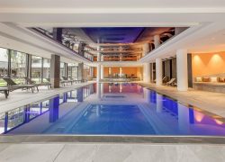 Vue générale d'une piscine intérieure d'hôtel pour un séminaire bien-être, comprenant une plage carrelée au premier plan, un bassin rectangulaire au centre, des chaises longues disposées à gauche et à droite, de grandes baies vitrées sur la gauche, et des colonnes blanches avec un plafond miroir en arrière-plan.
