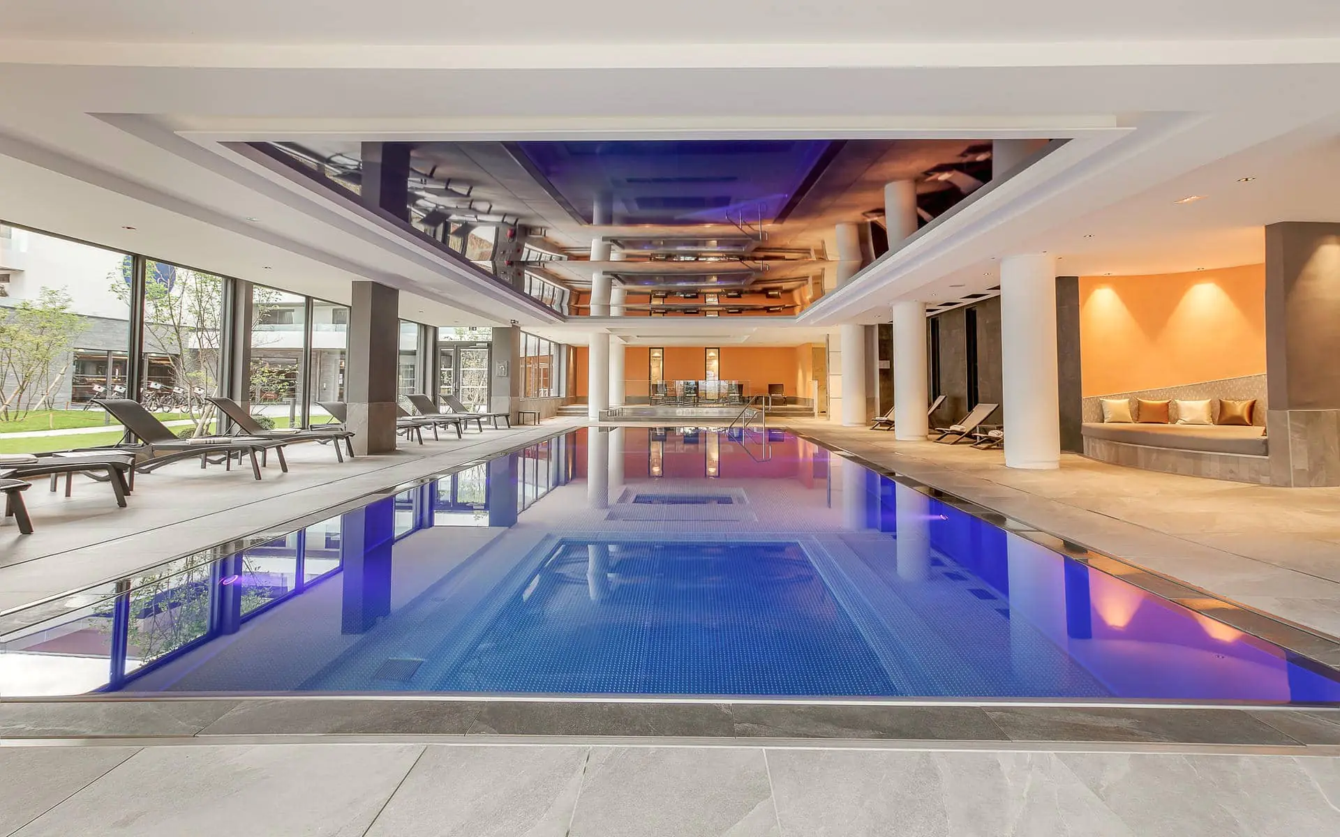 General view of an indoor hotel swimming pool for a wellness seminar, featuring a tiled deck in the foreground, a rectangular pool in the centre, sun loungers along the left and right sides, large floor-to-ceiling windows on the left, and white columns with a mirrored ceiling in the background.