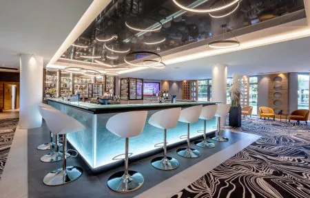 Hotel bar area for afterwork, with an illuminated central bar surrounded by white high stools in the foreground, bottle shelves and beer taps in the centre, patterned black and white carpet on the right, and large windows with lounge armchairs in the background.