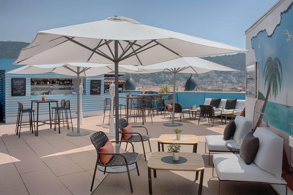Hotel rooftop terrace with large white parasols shading dark metal tables and chairs with terracotta cushions in the foreground, a light grey sofa with dark cushions on the right beside a wall painted with a palm tree and beach mural, a striped blue and white cocktail bar with high stools on the left, and hills with city rooftops visible in the background, a rooftop setup suited for an afterwork.