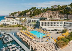 Vue aérienne d'un complexe hôtelier incarnant une destination séminaire en bord de mer, comprenant une piscine rectangulaire turquoise entourée de parasols jaunes et de chaises longues au premier plan, des bâtiments d'hôtel blancs à plusieurs étages et terrasses en toiture au centre, et un littoral rocheux avec port de plaisance et bateaux amarrés devant une falaise en arrière-plan.