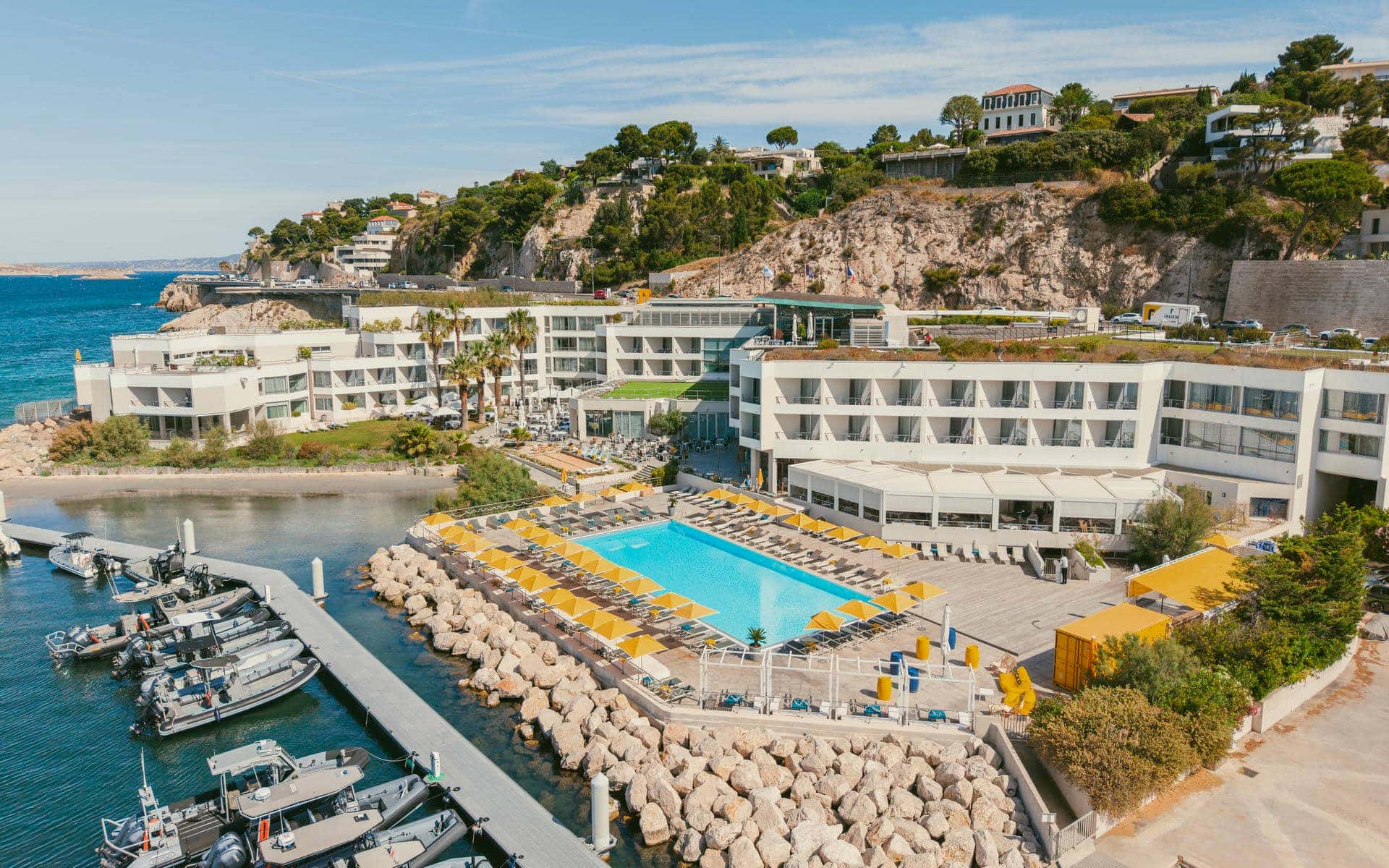 Vue aérienne d'un complexe hôtelier incarnant une destination séminaire en bord de mer, comprenant une piscine rectangulaire turquoise entourée de parasols jaunes et de chaises longues au premier plan, des bâtiments d'hôtel blancs à plusieurs étages et terrasses en toiture au centre, et un littoral rocheux avec port de plaisance et bateaux amarrés devant une falaise en arrière-plan.